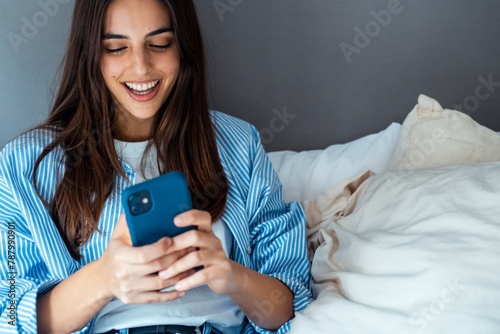 Young woman sitting on bed, dressed in casual clothes, holding phone and reading new messages, smiling peacefully. Technology concept.