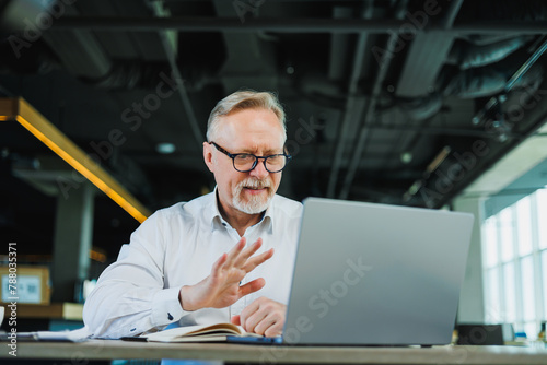 An adult bearded man is sitting at a laptop and working in the office. A gray-haired man runs an online business. photo