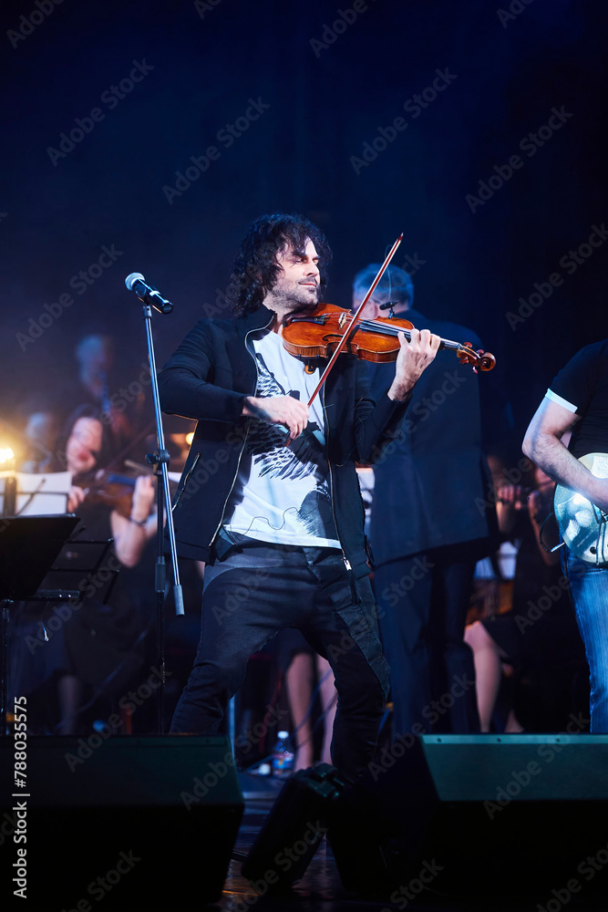 Fototapeta premium A violinist musician stands on stage with a violin in his hands during a concert. There is a bright light from the floodlights around.