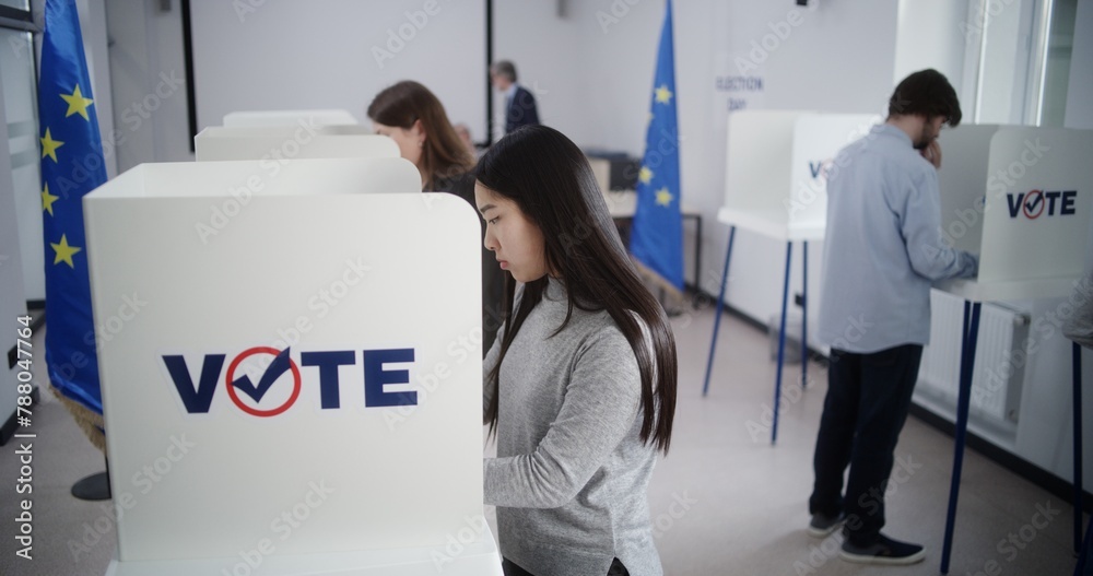 Obraz premium High angle of multiethnic European people voting at polling station during EU elections. Diverse voters fill out ballots in voting booths. Election Day in the European Union. Civic duty and democracy.