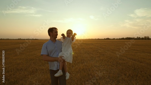 Father holding kid in arms in field at sunset. Happy cheery parent child kid strolling moving going across huge yellow meadow. Girl sits in man arms pointing with finger. Love fatherhood parenthood