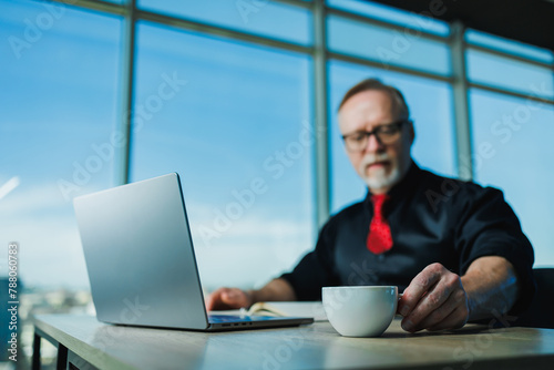 Adult gray-haired businessman sitting in office at workplace, old businessman in shirt working with laptop. A gray-haired businessman is sitting at the workplace.