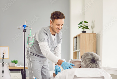 Male nurse helping and taking care of aged patient. Happy friendly man in scrubs and latex gloves standing by hospital bed with old diseased pensioner lady receiving liquid IV vitamin therapy solution