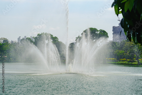 High jets of water from a fountain in a lake, surrounded by lush greenery in an urban park, under a clear sky. © InfinitePhoto