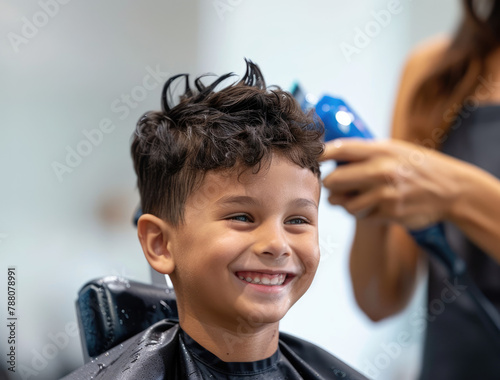 a boy getting a haircut in a hair salon, with a white wall and a black apron on the barber behind him. The happy child has his eyes closed and is sitting in the chair smiling