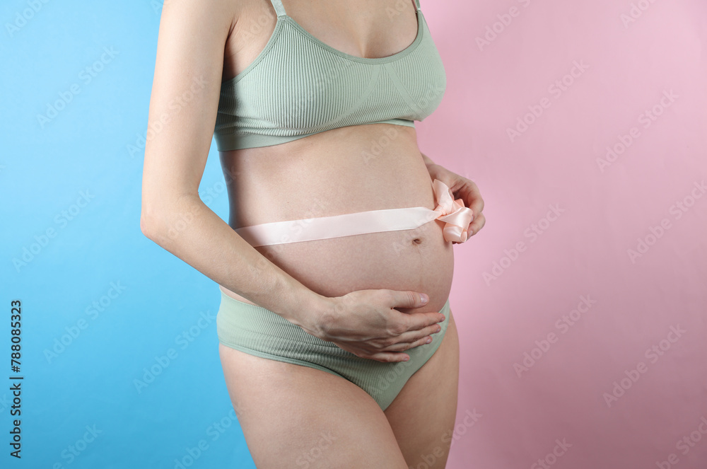 Pregnant woman in underwear with her belly tied in a bow on a blue-pink background Pregnant woman in underwear with her belly tied in a bow on a blue-pink background