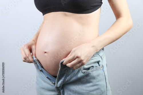 Pregnant woman putting on jeans on gray background