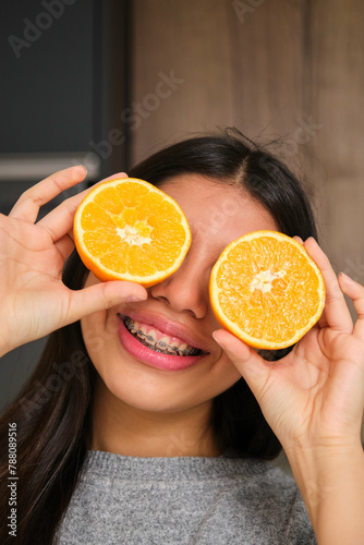 Happy female teenager with braces covering her eyes with an orange fruit. © Ladanifer