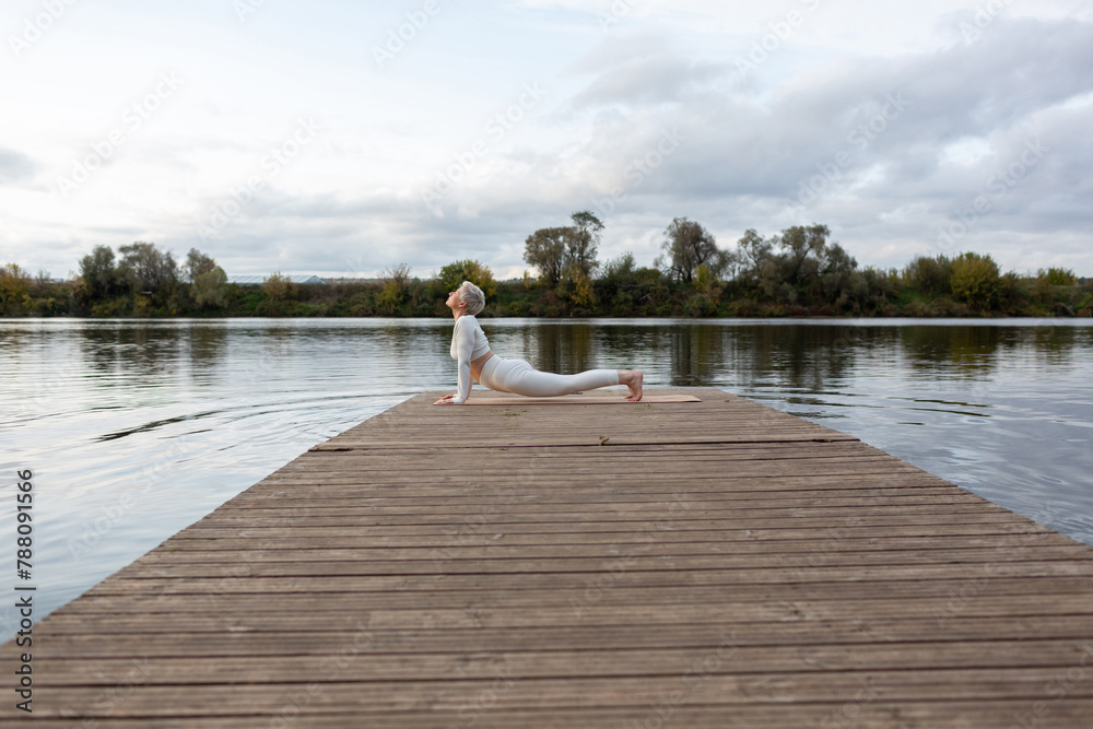 Fototapeta premium A mature woman with a short haircut in a white suit is doing yoga on the riverbank. Bhujangasana, Upward Dog