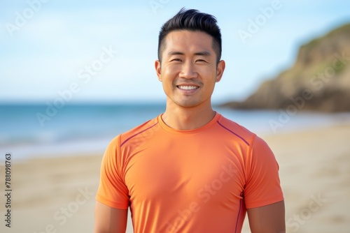 Portrait of a glad asian man in his 30s wearing a moisture-wicking running shirt while standing against sandy beach background © Markus Schröder