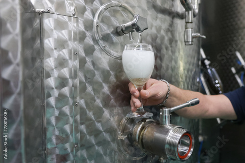 Details with the hand of a winemaker pouring sparkling wine from a metal tank into a glass, in a modern winery.
