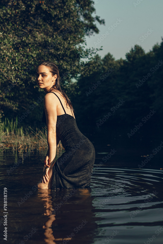 Sexy graceful woman with wet hair in dress standing in water of lake in summer and looking at camera 