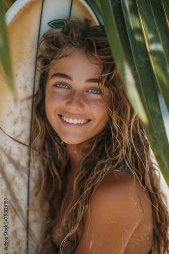  beautiful hot young surfer woman with wet wavy blonde hair, charming smile , piercing blue eyes, sensitive gaze, tanned skin . she is holding a serf board . summer beach setting,tropical leaves