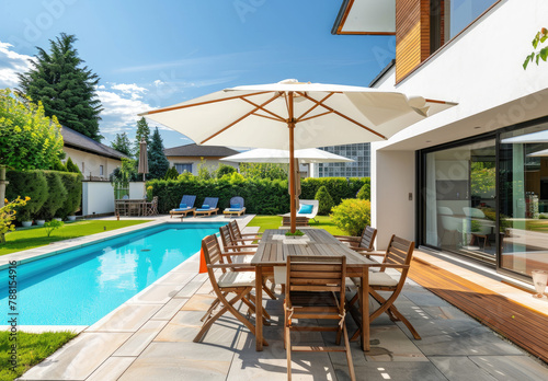 Modern garden with a swimming pool, wooden table and chairs under an umbrella on a sunny summer day next to a modern house with glass windows. © Kien
