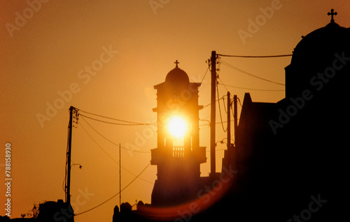 Unter-oder aufgehende Sonne leuchtet orange durch den Glockenstuhl einer Kirche auf der griechischen Insel San Turin photo
