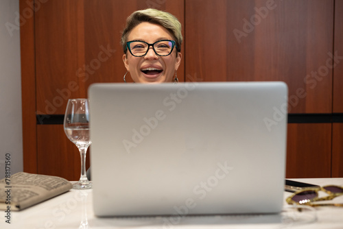 a girl with short hair and glasses indoors at a laptop laughs and smiles emotionally and cheerfully while working and chatting