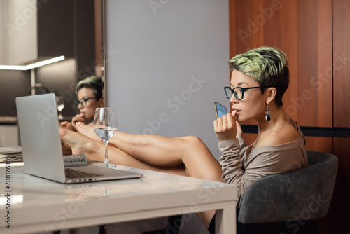 a beautiful girl with short hair and glasses is sitting indoors at a laptop, with her feet on the table, chatting and working online