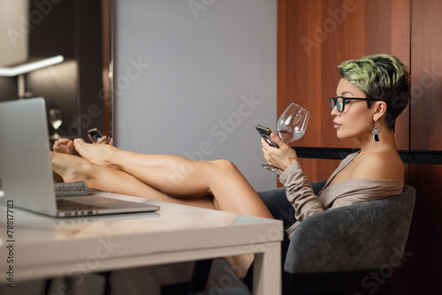 a beautiful girl with short hair and glasses is sitting indoors at a laptop, with her feet on the table, chatting and working online with a glass of water