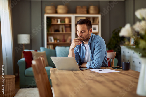 A focused male remote worker resting his hand on his chin and revising his work on a laptop. photo