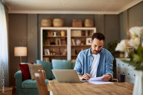 A focused young adult male freelancer reading a document in front of him while sitting in front of a laptop.