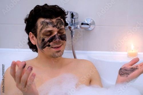 Man with charcoal face mask lying in bath with foam and candle meditating at home. pa, hygiene, skin care procedure, treatment, wellness. Handsome young brunette male relaxing in bathroom in evening. photo