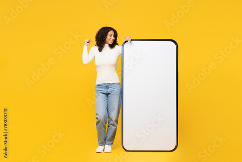 Full body little kid teen girl wear white casual clothes big huge blank screen mobile cell phone smartphone with workspace area do winner gesture isolated on plain yellow background Lifestyle concept