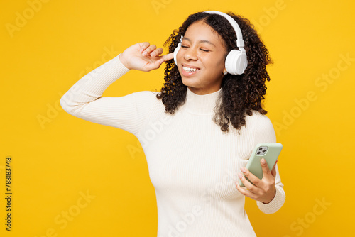 Little kid teen girl of African American ethnicity wear white casual clothes listen to music in headphones use mobile cell phone isolated on plain yellow background studio Childhood lifestyle concept photo