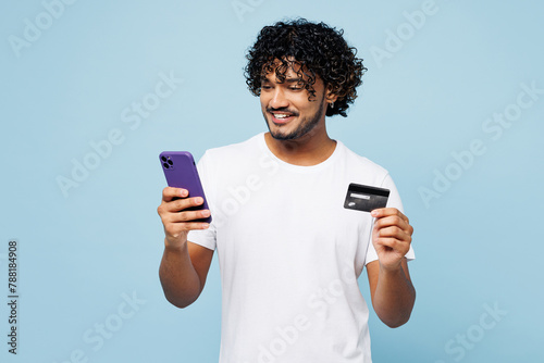 Young Indian man wear white t-shirt casual clothes using mobile cell phone hold credit bank card do online shopping order delivery book tour isolated on plain blue cyan background. Lifestyle concept.