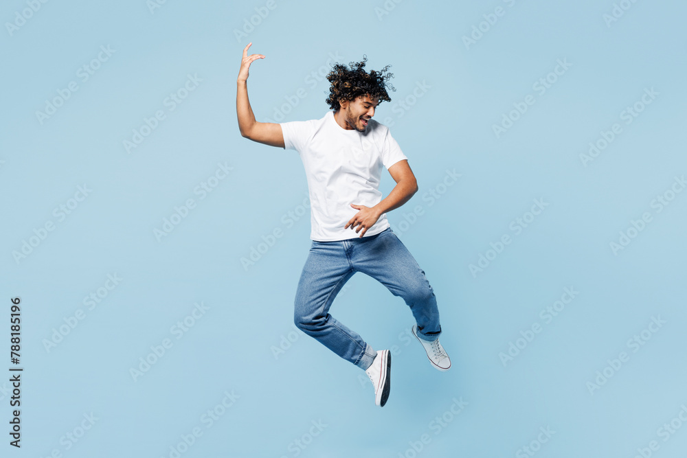 Full body young happy fun Indian man he wears white t-shirt casual clothes jump high play air guitar jump high isolated on plain pastel light blue cyan background studio portrait. Lifestyle concept.