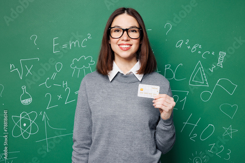 Young happy smart teacher woman wear grey casual shirt glasses hold credit bank card isolated on plain green wall white chalk blackboard background studio. Education in high school college concept. photo