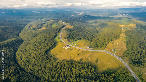 Vertical drone view above a mountain pass, crossing a serene meadow with coniferous forests on it. An asphalt road winds along the mountain peaks under the horizon. Sunset time.
