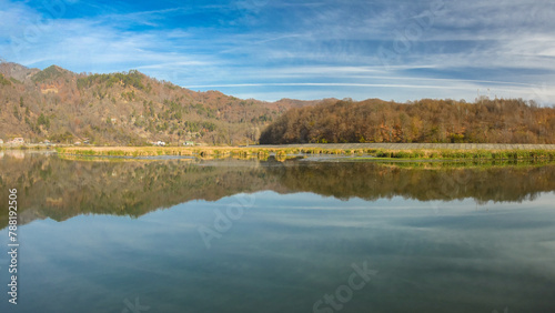 A quiet lake with reeds and swamps narrowing its shores. The lake is located in a rural area, surrounded by hills and beech forests. The sky is mirroring in its waters. Olt Valley, Carpathia, Romania.