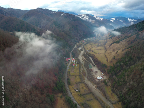 Aerial drone panorama above a valley inside a hilly region. Winter season, the mountains are snowed. The rural area is covered with low altitude clouds that mix with the beech forests. Carpathia,