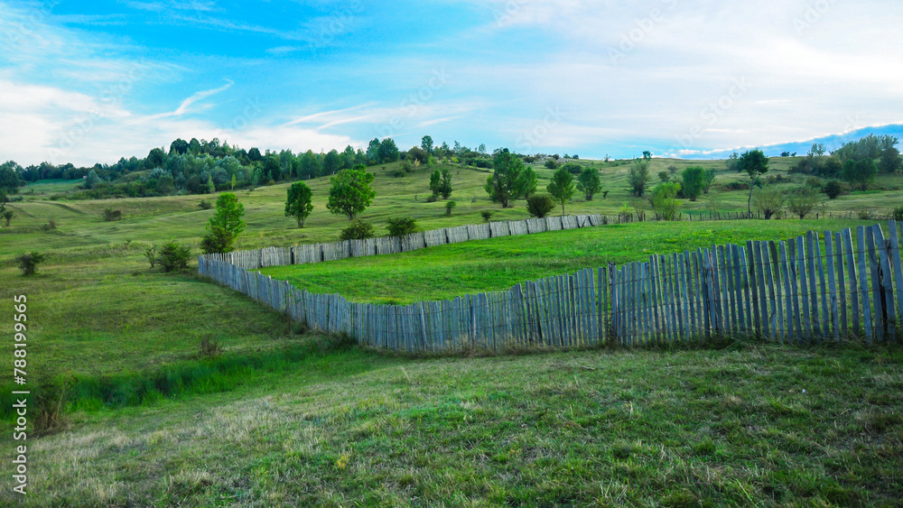 A wooden fence built on a hill. The meadows on the hill are used for farming or for orchards. Rural landscape aof traditional farming in Eastern Europe. 