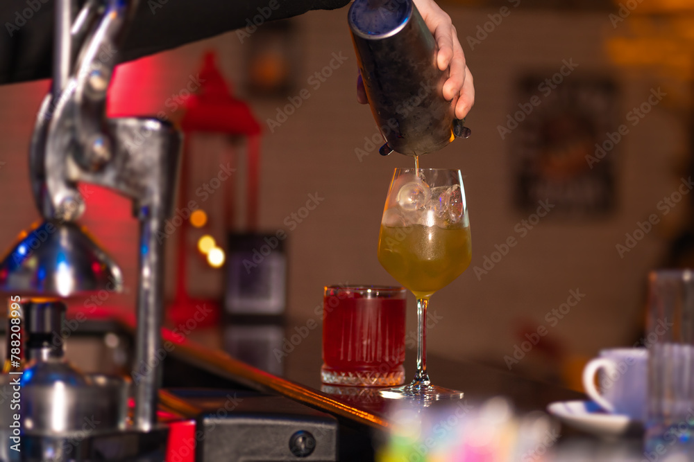 Close-up of the bartender carefully pours a ready-made cocktail from a metal shaker into a glass with ice behind the bar. - obrazy, fototapety, plakaty 