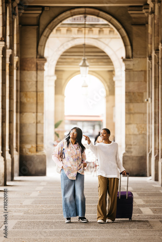 Obraz na płótnie Two young cheerful women pulling a suitcase and walking together through a city during a weekend trip