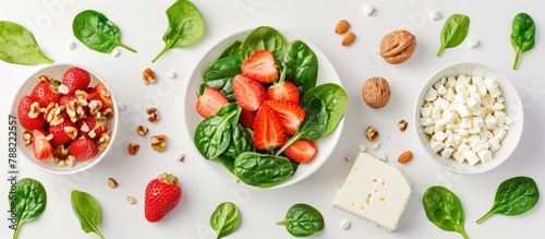 A strawberry salad with spinach leaves, sliced strawberries, nuts, and feta cheese laid out on a white background. Depicting the concept of healthy food in a flat lay, top-down view.