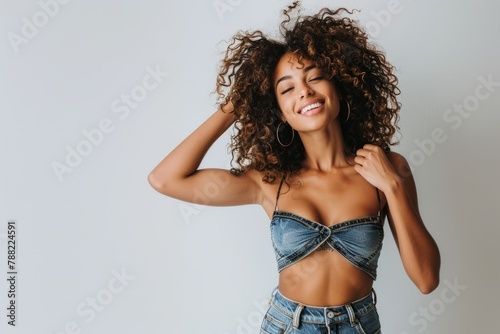  A beautiful mixed race model with curly hair, smiling and posing in front of the camera wearing a denim vest top, hands behind her head against a white background in a studio photography setting with