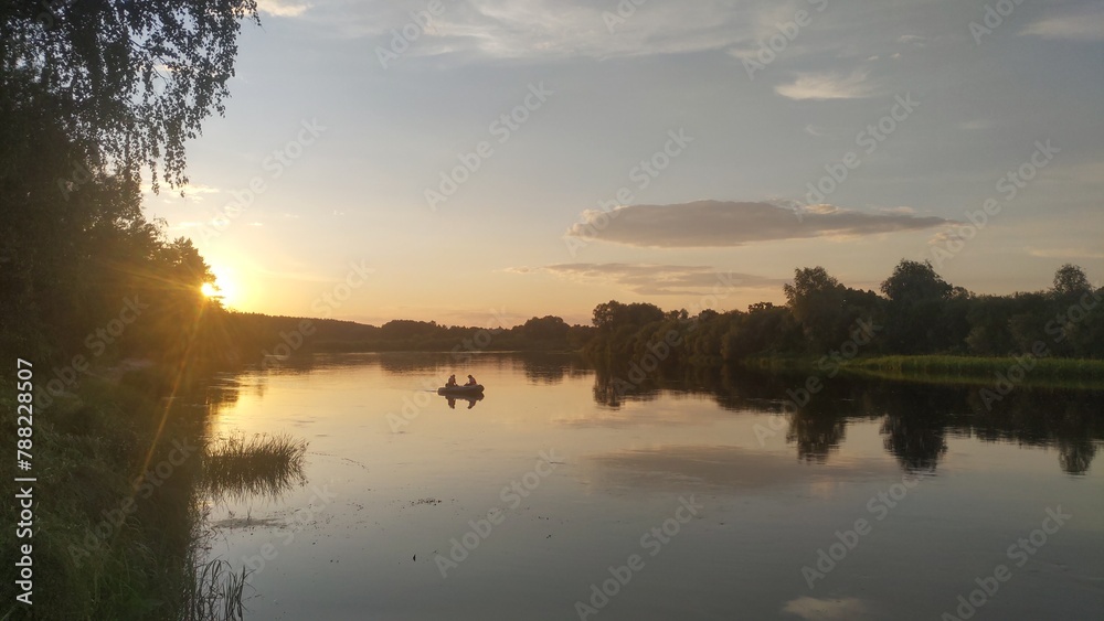 A PVC motorized inflatable boat floats on the river, while fishermen catch predatory fish by trolling. Illuminated by the summer sunset sun, tree branches lean over the water. Trees and shrubs grow