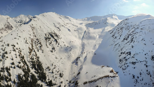Aerial drone panorama above Fagaras mountain peaks. Springtime season, the crests are covered with deep snow. Several sprice trees grow on the mountainsides. Carpathia, Romania.