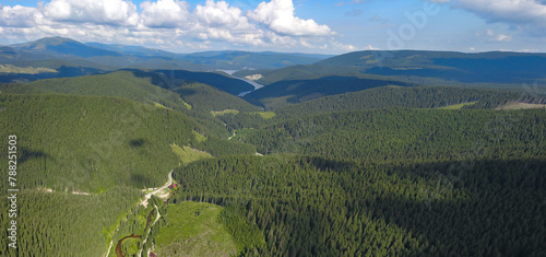 Aerial drone view above Oasa Lake and Sureanu Massif with its high altitude peaks and vast coniferous forests. Below, Transalpina road winds through the valley., along the river. Carpathia, Romania.