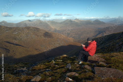 Hiker man sitting on the big stone on the top of the mountain and using his phone at the background of morning landscape with mountains. View from The Devils Staircase to Binnein Mor. Scotland