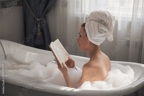 Back view of beautiful young woman reading book while enjoying bubble bath. Lady on hotel vacation lying in relaxing bathtub with paper book. Young lady reading book while relaxing in float bath