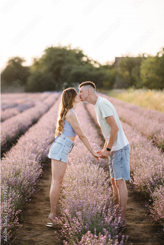 Young couple holding hands and kissing together in purple lavender field. Male and female enjoying romantic time in sunset. Beautiful woman and man walking among violet flowers with sunlight on summer