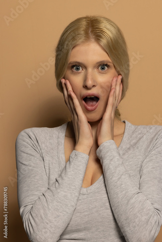 Portrait of beautiful young shocked woman with long tied blond hair on beige background