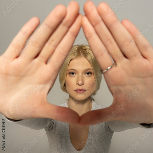 Attractive young woman looking trough the fingers frame on gray background