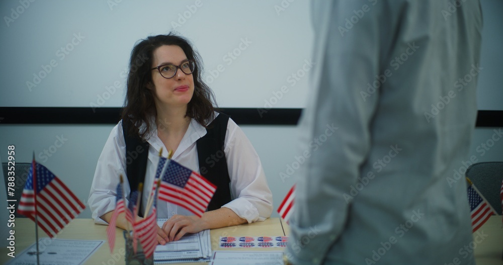 Female polling officer sits at the registration table at polling station, talks with African American male voter and gives paper bulletin. Election Day in the United States. Civic duty and democracy. Female polling officer sits at the registration table at polling station, talks with African American male voter and gives paper bulletin. Election Day in the United States. Civic duty and democracy.