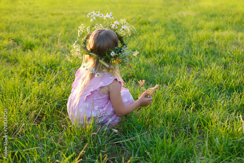 floral crown, wreath adorns head child, happy girl holding chamomile flower in hand on green sunlit meadow, happy childhood, natural world, seen from behind, Midsummer celebration