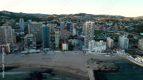 Sunset over famous city of Calp. Sunlight reflecting on skyscrapers and landscape. Drone going backwards above the bay. View of Cantal Roig Beach and promenade of the city. Travel destination photo
