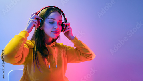 Portrait of a calm Caucasian woman wearing a yellow hoodie sitting and wearing wireless headphones, and posing isolated on a neon light background. Enjoying new albums of favorite music and artists.